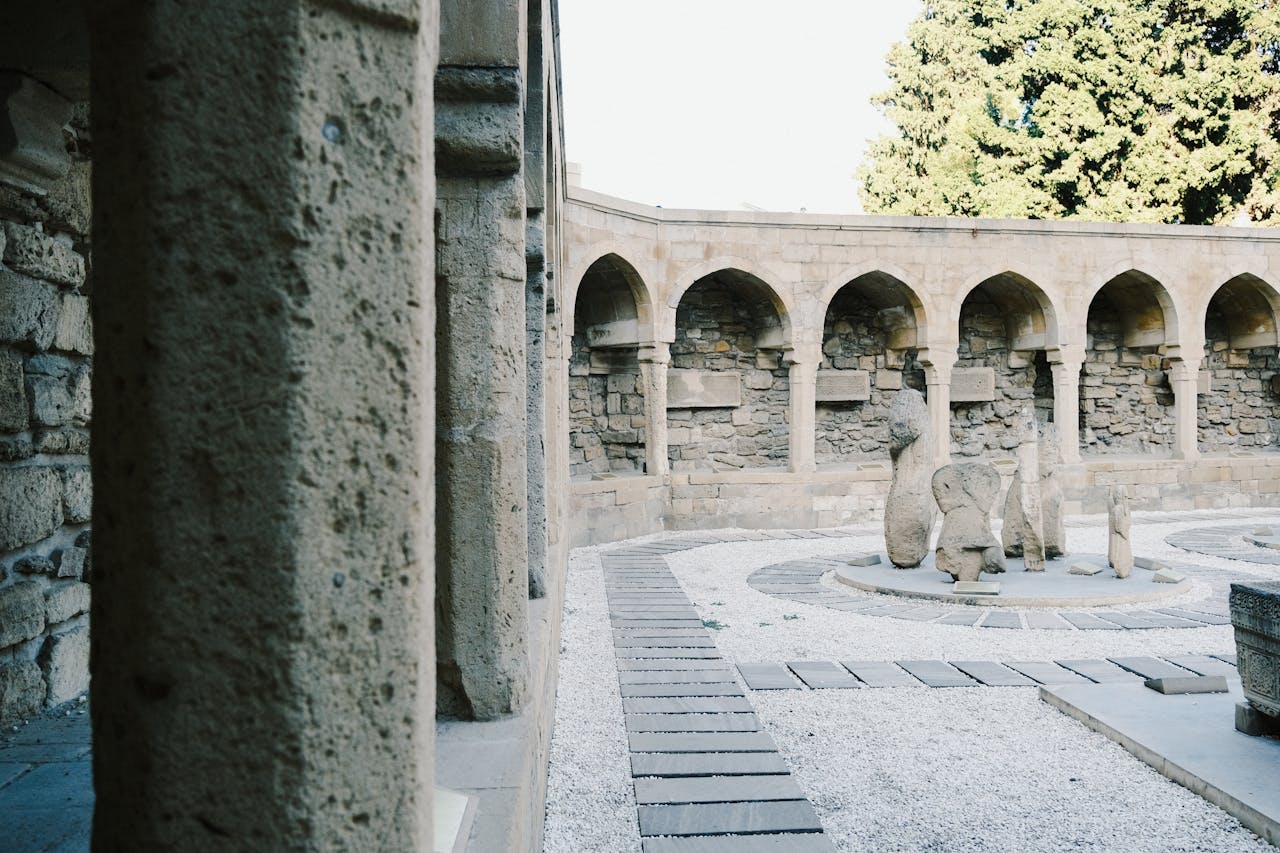 Stone courtyard with arches and sculptures, bathed in sunlight, surrounded by trees.
